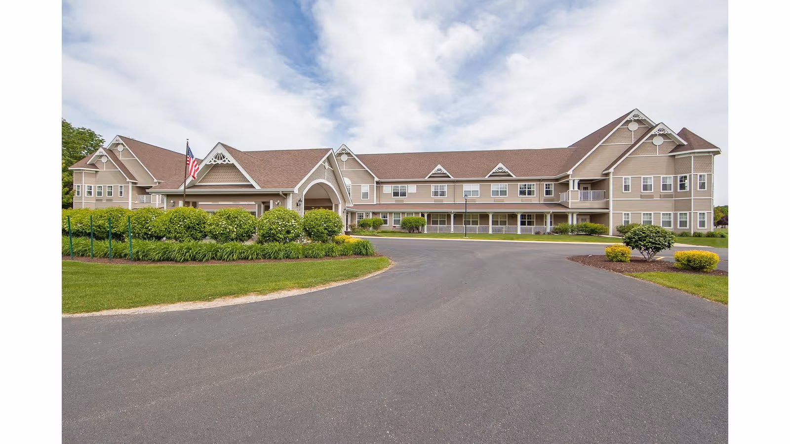 Exterior front view of a large two-story senior living facility building with beige siding and a brown roof. The building features multiple windows, decorative gables, and a covered entrance with an American flag. The surrounding area includes a paved driveway, green lawns, bushes, and small landscaped areas under a partly cloudy sky.