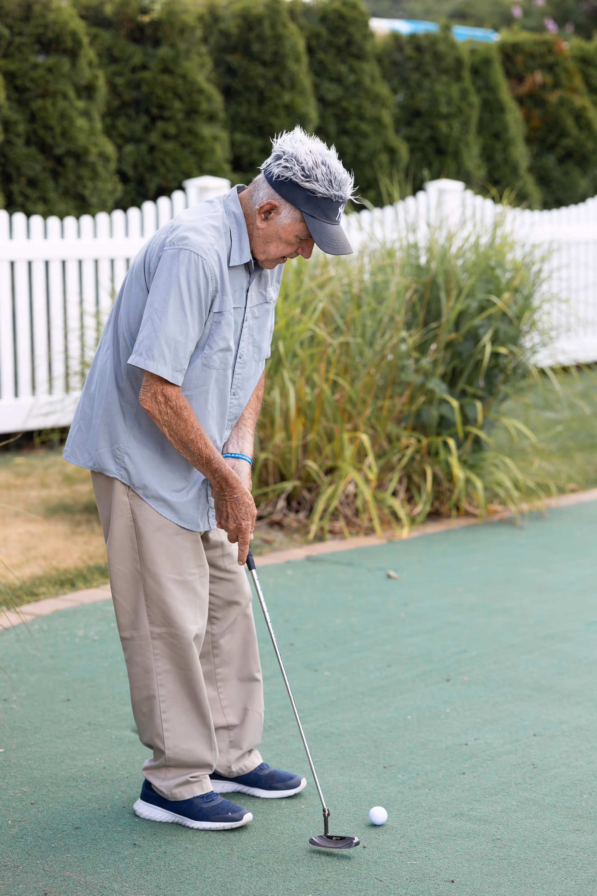 An elderly man wearing a gray shirt, beige pants, and a black visor is playing mini-golf on a green putting surface. He is focused on putting a golf ball with a putter. Behind him is a white picket fence and green bushes.