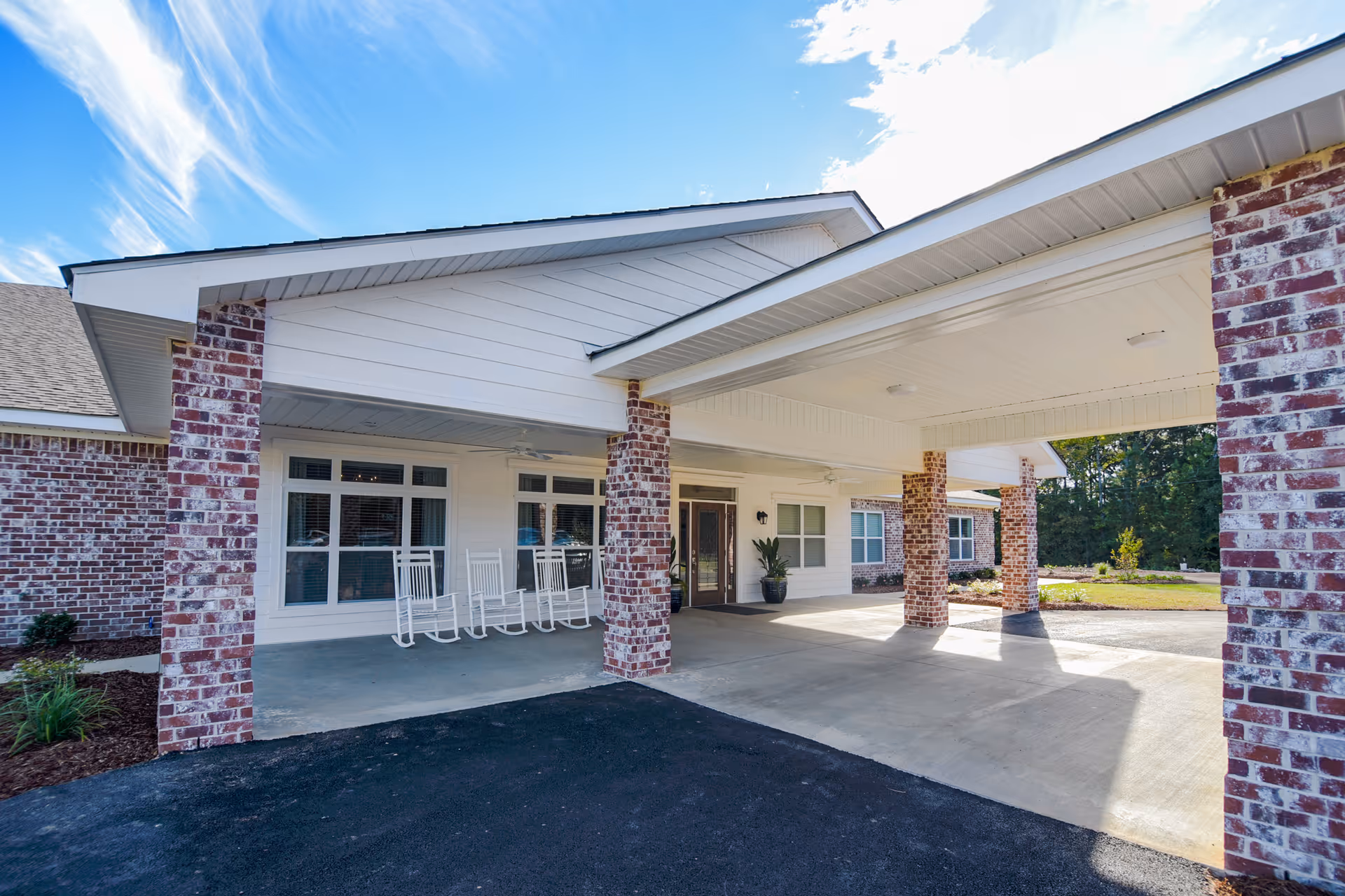 Covered entrance of a brick senior living building with rocking chairs on the porch and a drive-through porte-cochère.