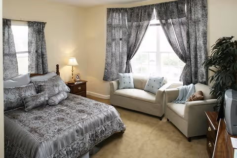 Cozy bedroom featuring a patterned bed, two windows with curtains, and a seating area.