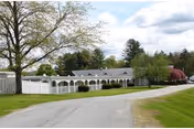Single-story nursing home with a white arched veranda, surrounded by green lawn and trees.