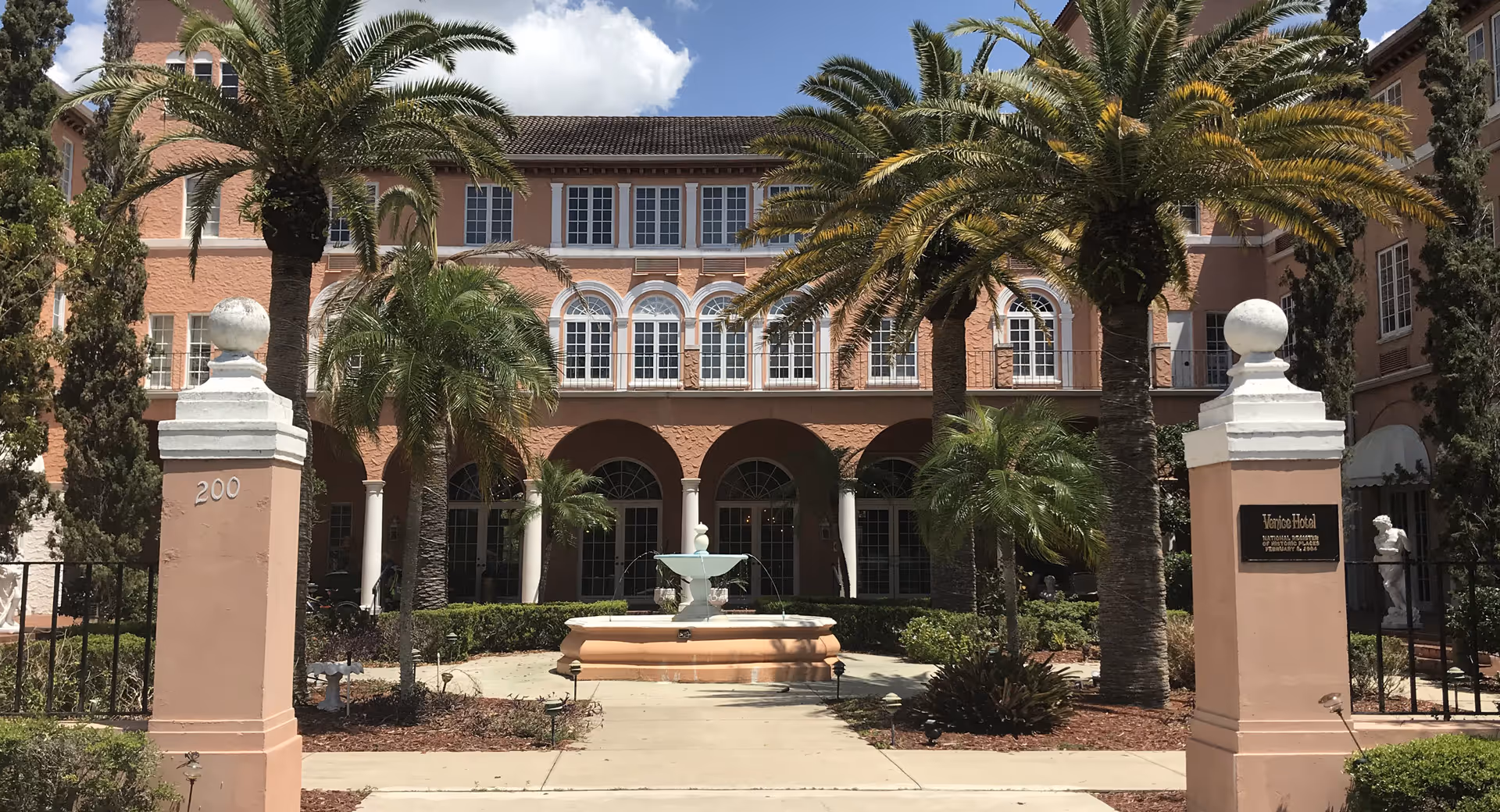 Exterior view of a peach-colored building with arched windows and columns, surrounded by palm trees and greenery. In front of the building is a circular fountain with water jets. Two large pillars mark the entrance, one with the number 200 and the other with a plaque that reads 'Venice Hotel National Register of Historic Places February 17, 1986.'