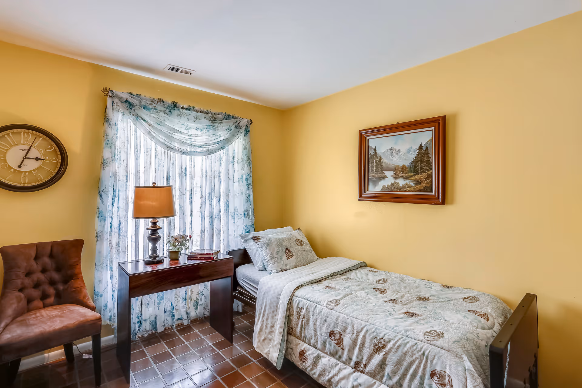 A small bedroom with yellow walls featuring a single bed with patterned bedding, a wooden side table with a lamp and small plant, a brown upholstered chair, a wall clock, and a window with sheer floral curtains. A framed landscape painting hangs on the wall above the bed.