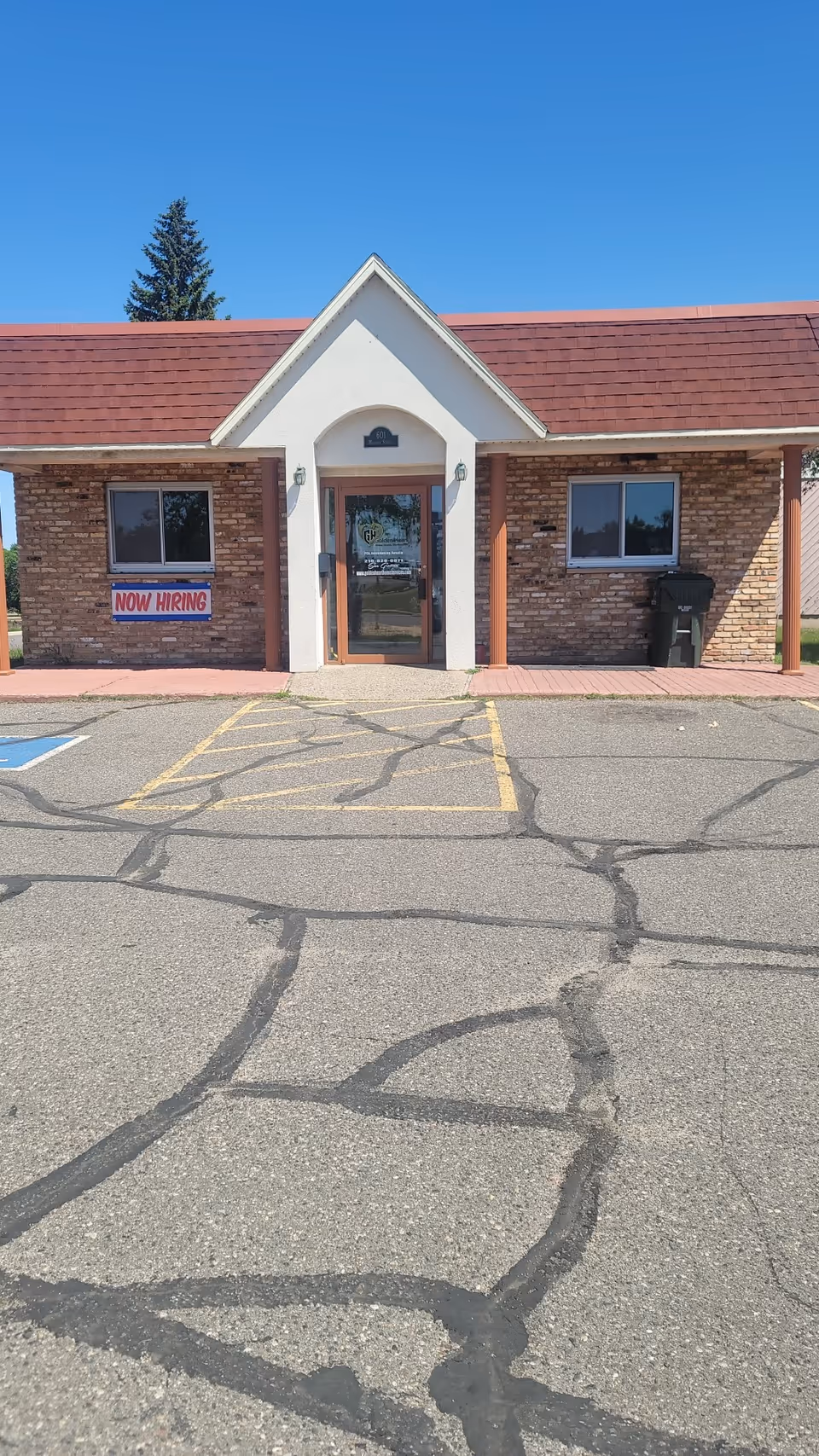 Exterior view of a brick healthcare facility with a central entry and a now hiring sign.