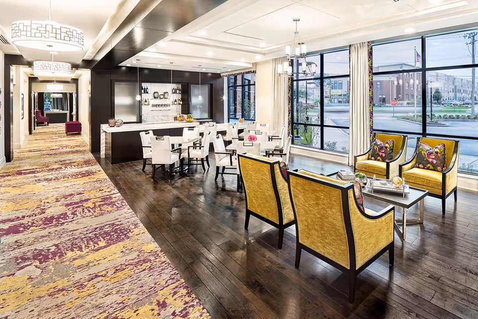 A bright and modern interior space featuring a seating area with four yellow armchairs around a coffee table, a dining area with several tables and white chairs, and a bar area with shelves holding bottles and glasses. Large windows with cream curtains provide a view of the outside buildings and street. The floor is dark wood with a patterned carpeted hallway on the left side, and contemporary light fixtures hang from the ceiling.