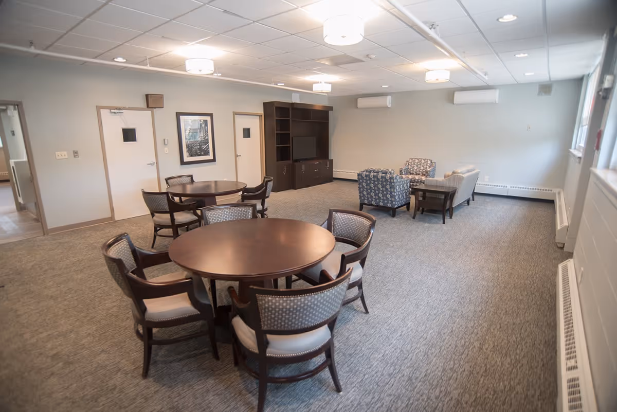 A spacious common area in a senior living facility with two round wooden tables surrounded by chairs in the foreground. In the background, there is a seating area with two patterned armchairs, a sofa, and a coffee table. A large dark wooden entertainment center with a TV is against the far wall. The room has carpeted floors, light gray walls, and ceiling lights.