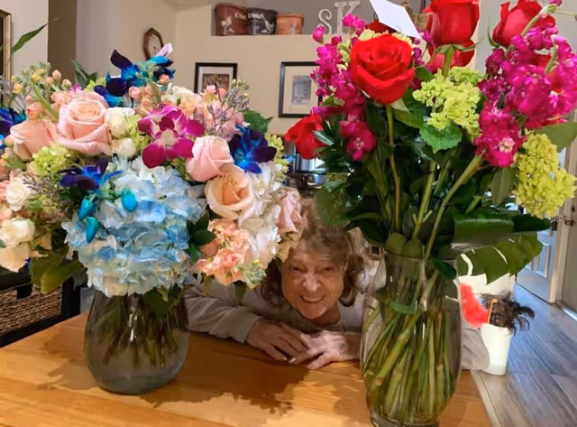 An elderly woman smiles behind two large bouquet arrangements on a wooden table indoors.