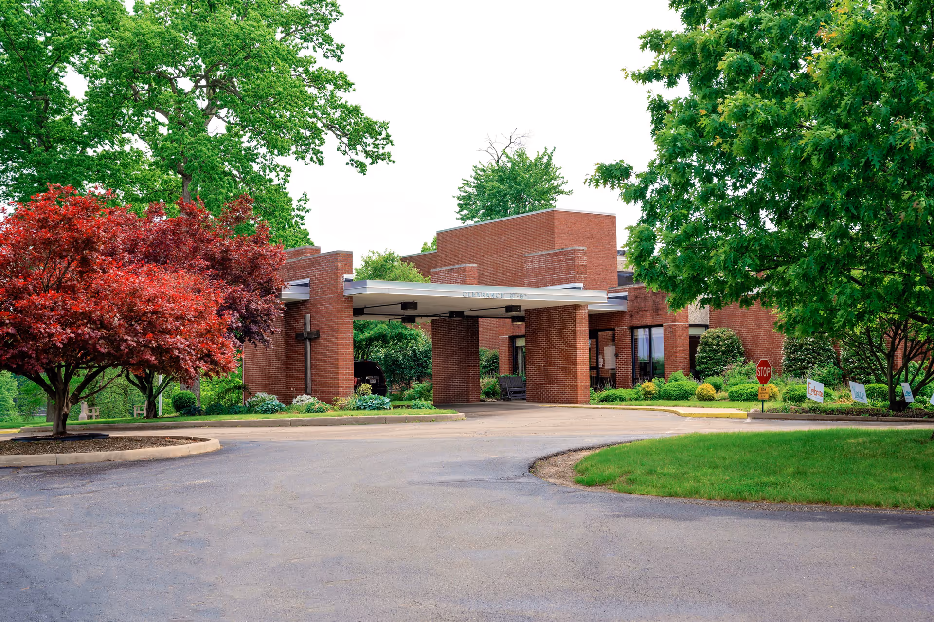 Exterior view of The Chapel Hill Community building with a covered entrance. The building is made of red brick and surrounded by green trees and landscaping, including a red-leafed tree on the left and a large green tree on the right. There is a paved driveway leading up to the entrance.