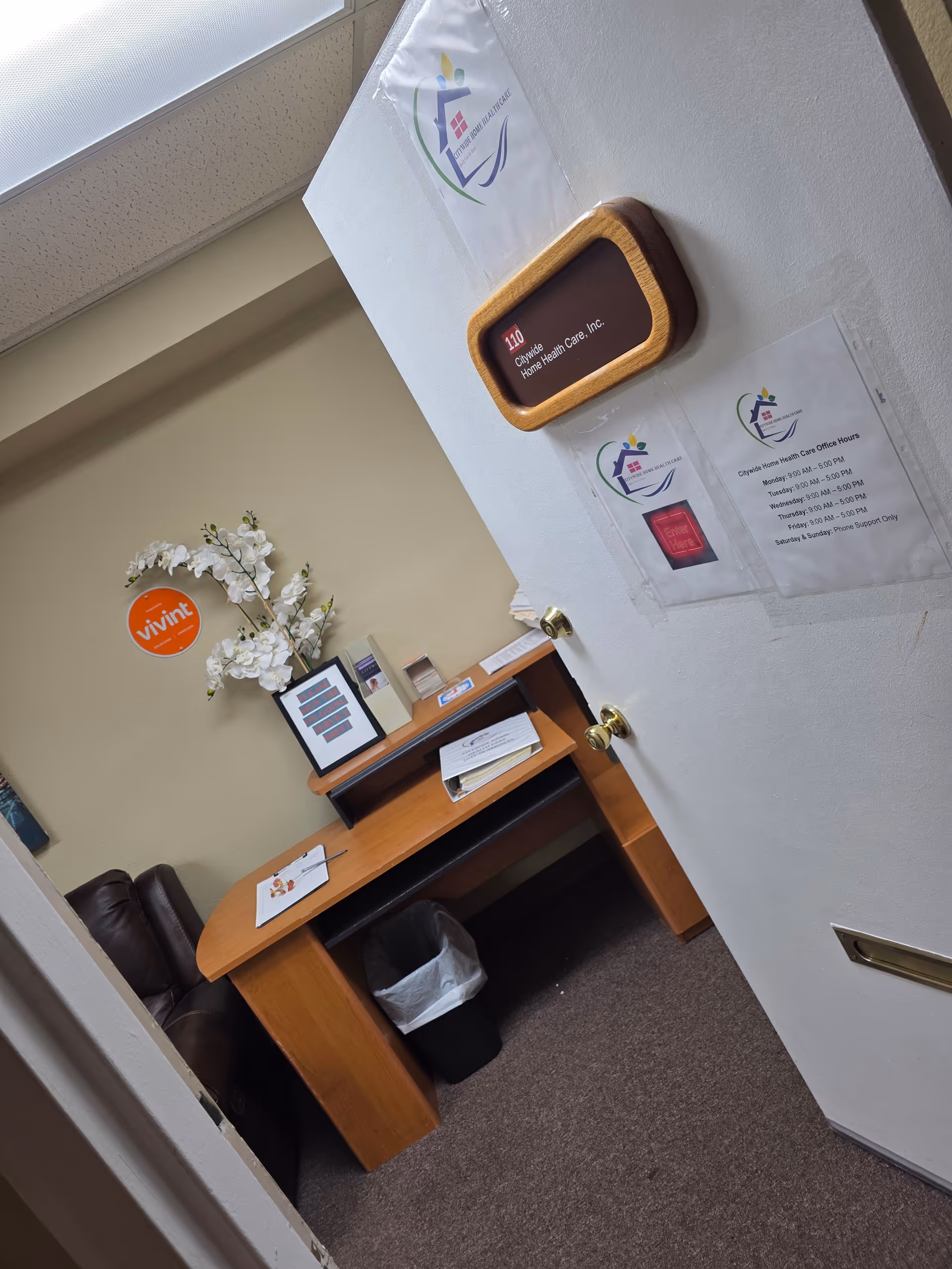 Interior office corner with wooden desk, paperwork, flowers, posters, and a trash can.