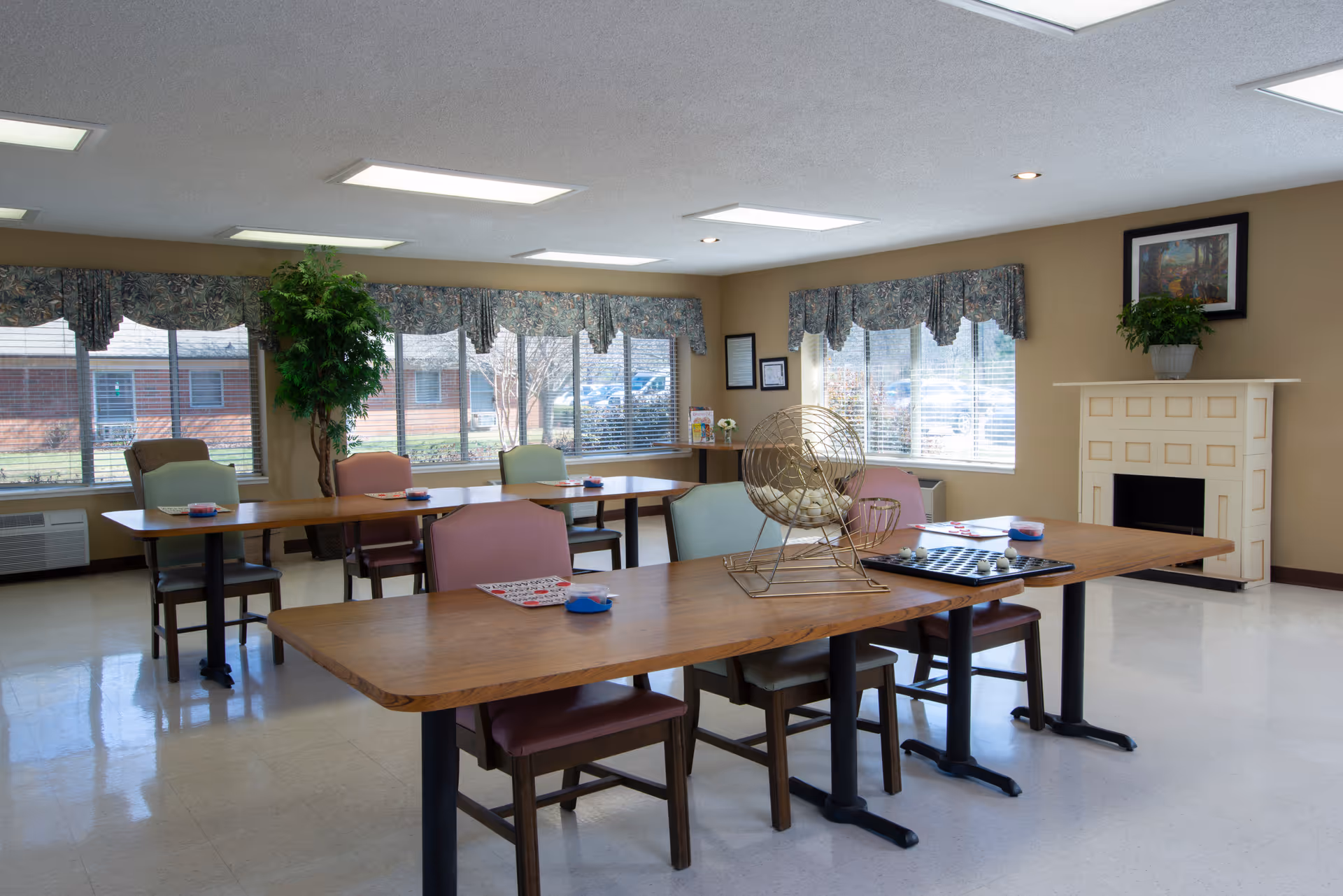 Bright senior living dining area with long wooden tables, chairs, windows, and a fireplace.
