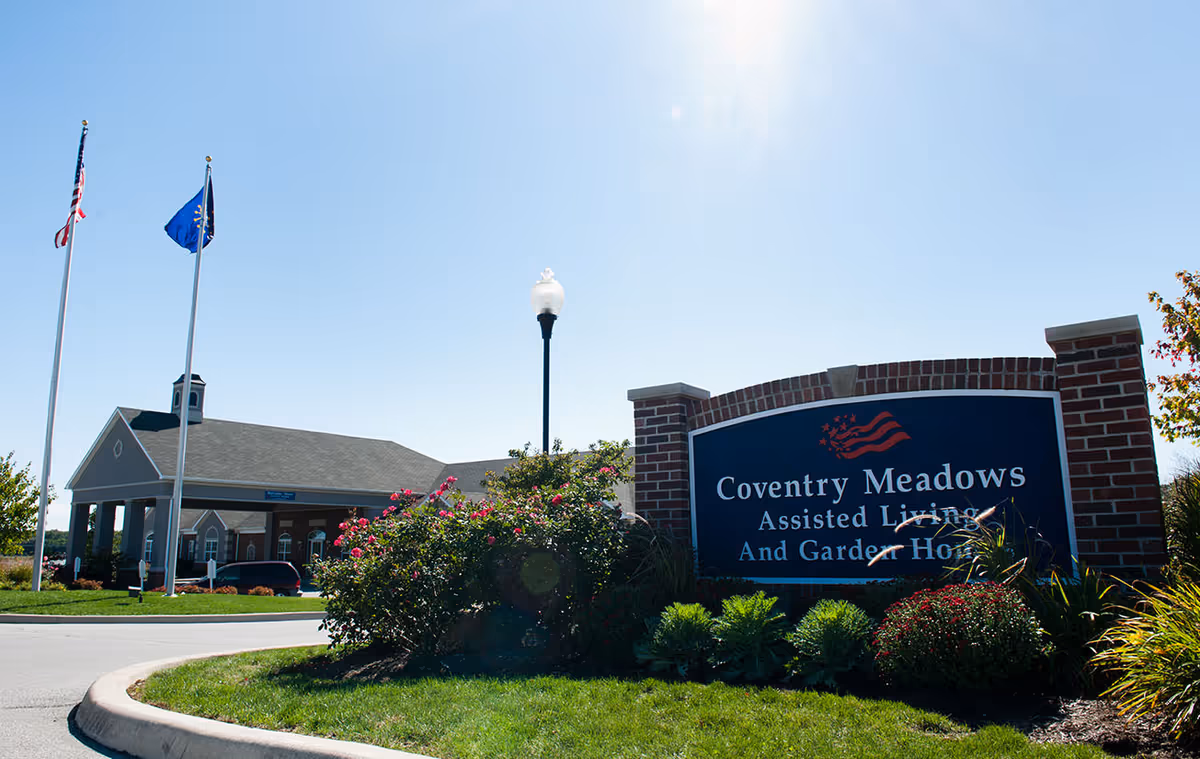 Exterior view of Coventry Meadows Assisted Living and Garden Home facility with a large brick sign in the foreground, two flagpoles with American and state flags, a street lamp, and a building entrance in the background under a clear blue sky.