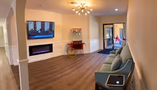 A cozy living room area with a modern wall-mounted flat screen TV above a sleek electric fireplace. There is a blue cushioned bench with pillows on the right side and a small table with a tablet on it. The room has wooden flooring and warm lighting from a ceiling fixture. In the background, there is a popcorn machine and a doorway leading to another room.