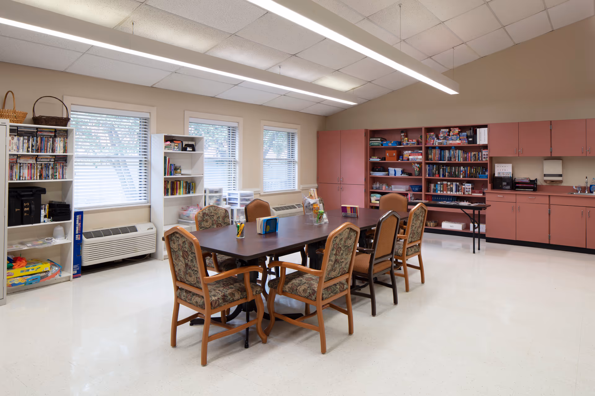 Community activity room with a large table, chairs, bookshelves, and pink cabinets.
