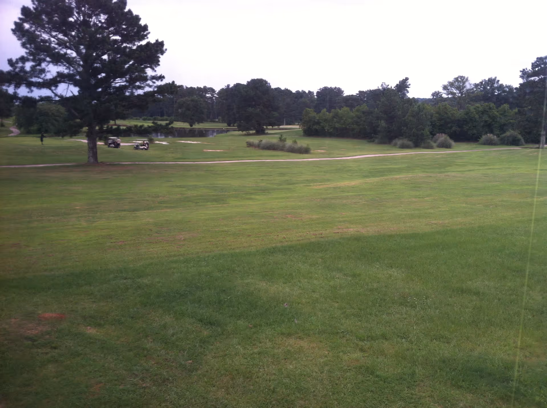 Wide view of a grassy golf course with trees, a pond, and two golf carts near a path.