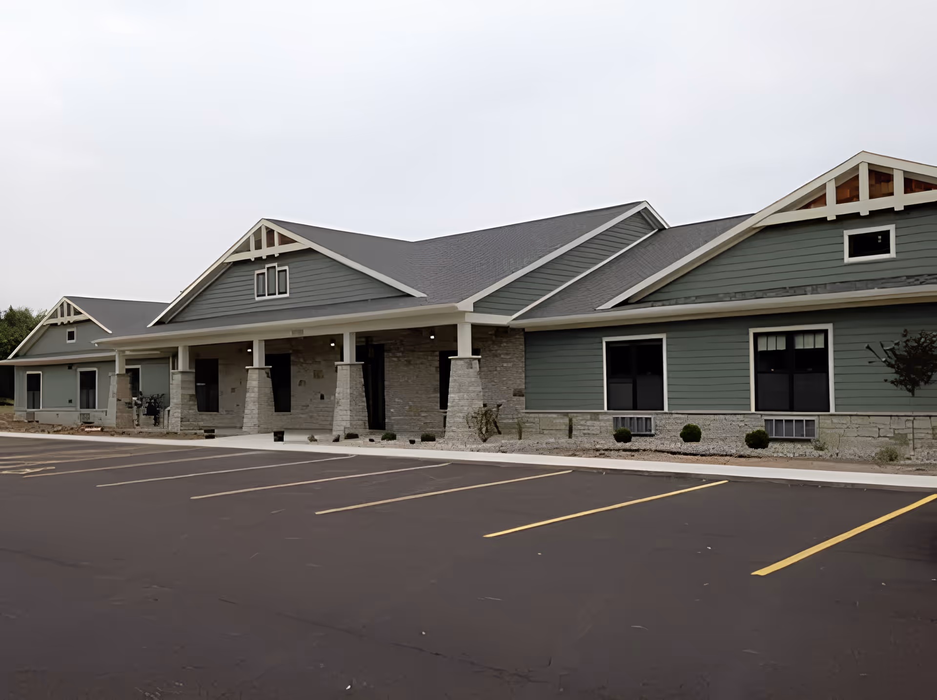 Single-story green and stone assisted living building with a covered entrance and empty parking lot in front.