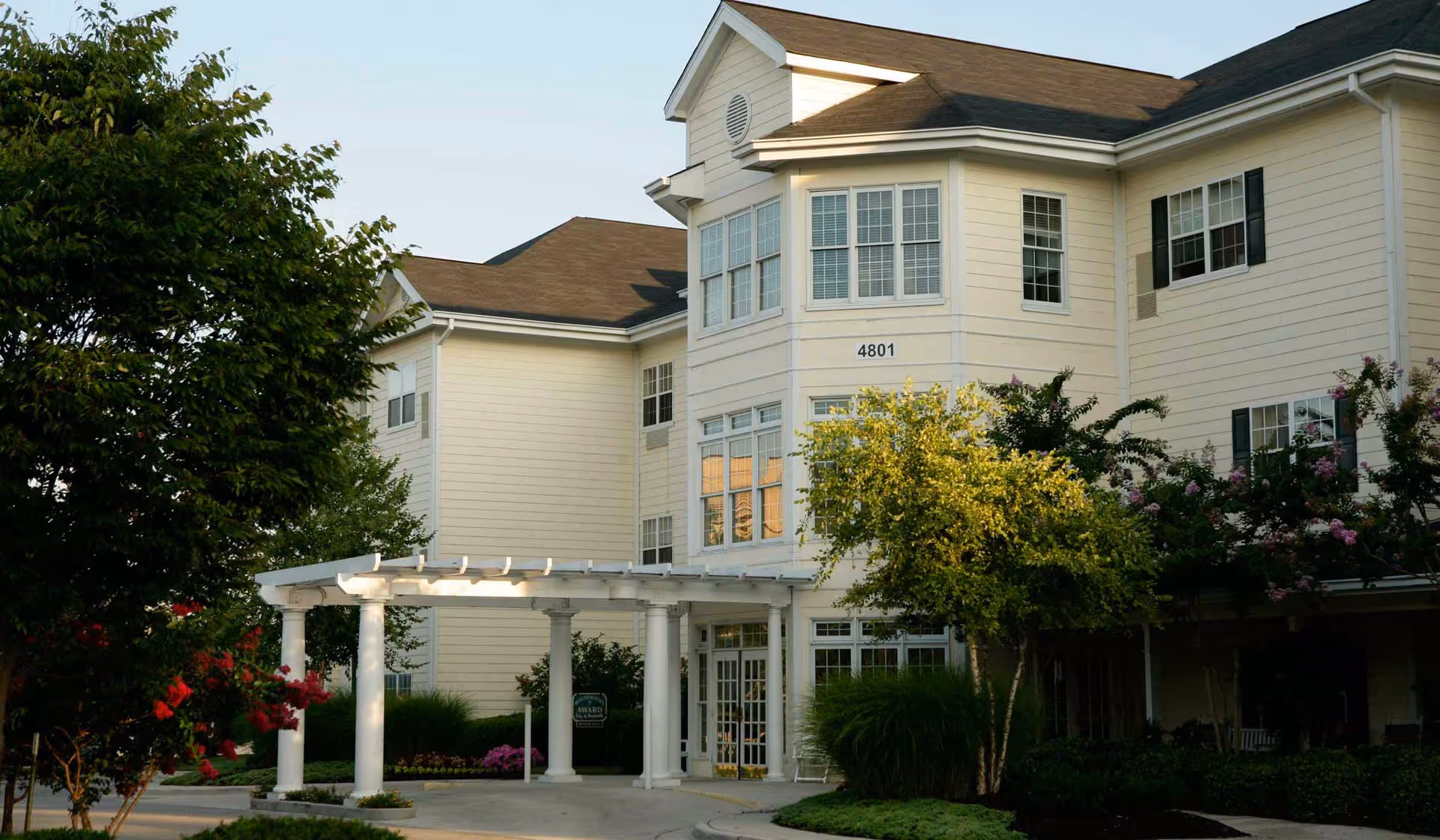 Cream-colored multi-story senior living building front with a covered white pergola entrance, trees, and landscaping.