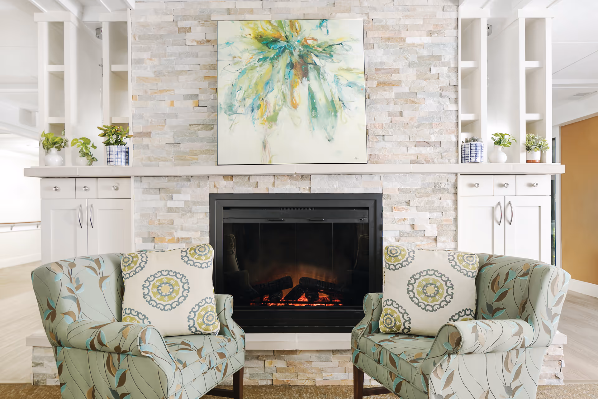 Stone fireplace with framed artwork and two patterned chairs in a bright living area.