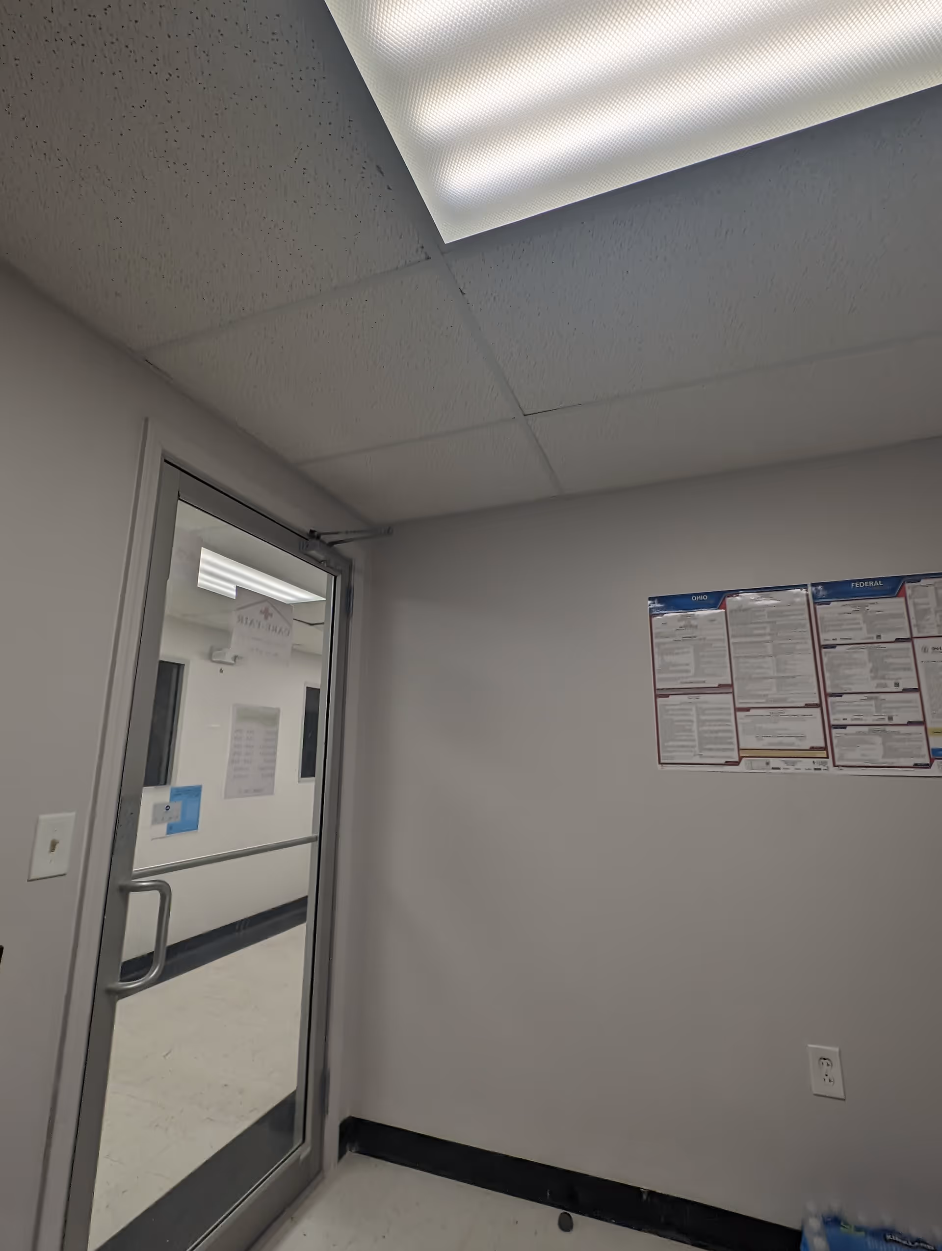 Interior hallway corner with a glass door, ceiling panels, and a bulletin board on the wall.