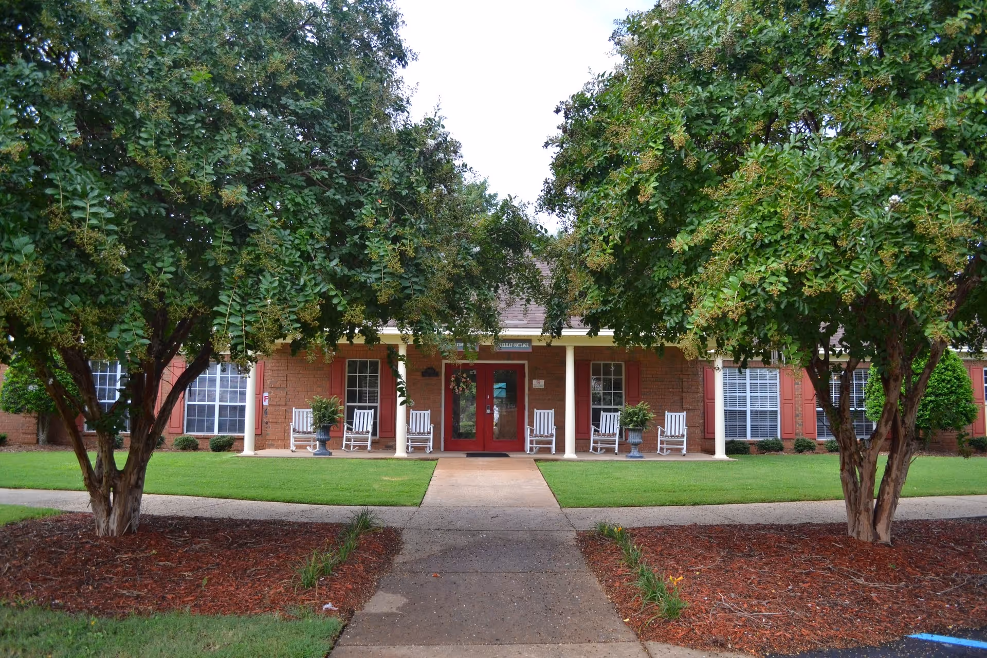 Front exterior view of a brick building with a covered porch featuring white rocking chairs and potted plants. The entrance has double red doors and is flanked by large windows with red shutters. Two large trees frame the walkway leading to the entrance.