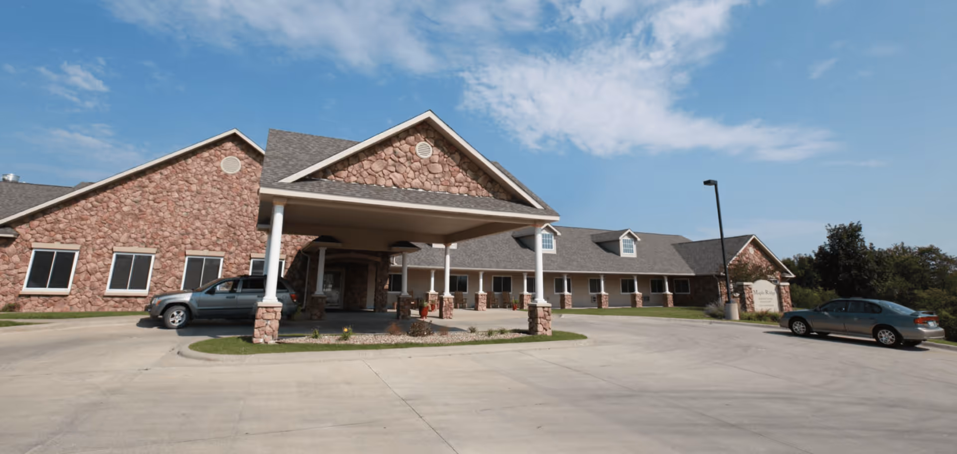 Exterior view of a single-story assisted living facility with stone and beige siding, a covered entrance supported by white columns, two parked cars, and a clear blue sky with some clouds.