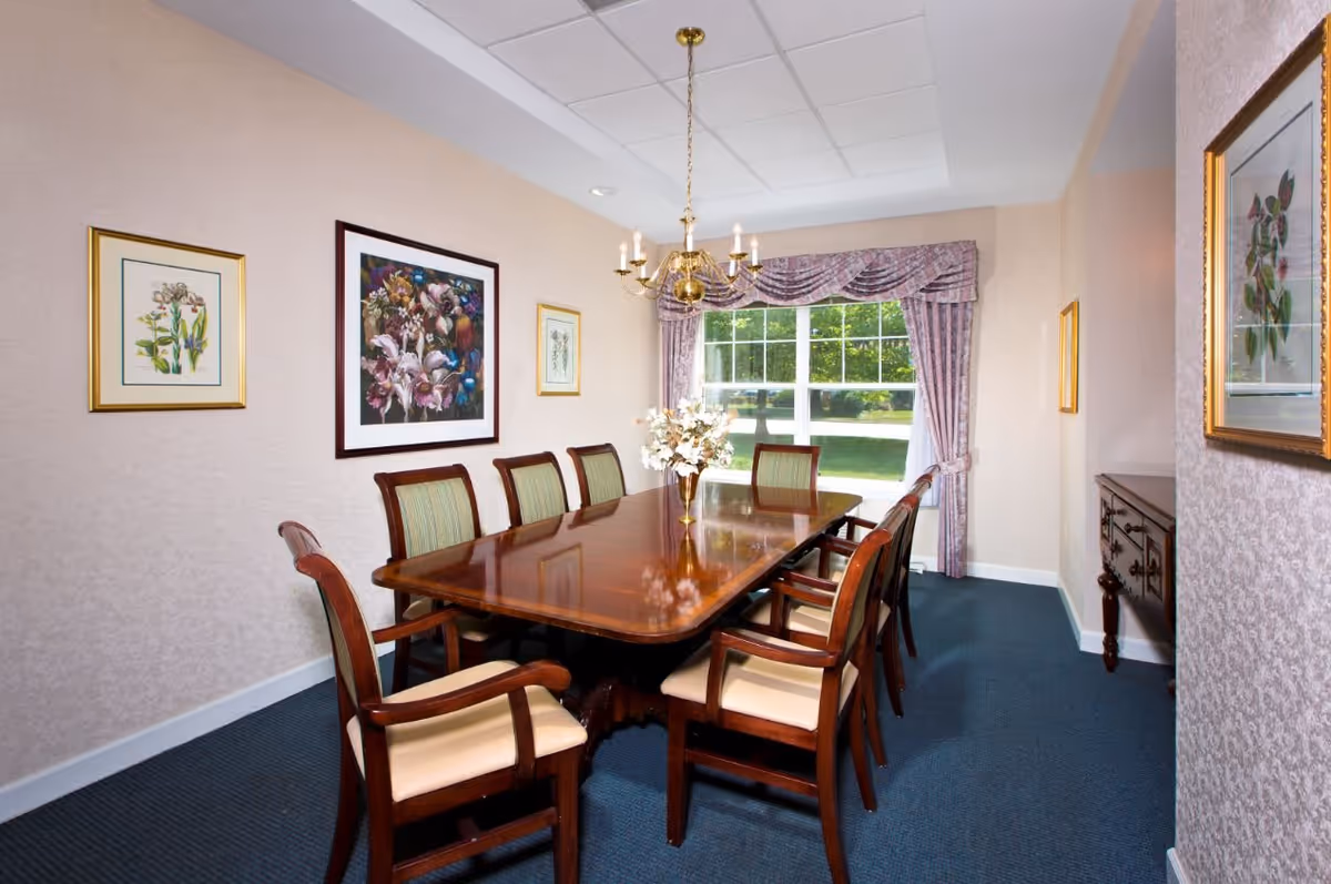 A formal dining room with a long polished wooden table surrounded by eight wooden chairs with cushioned seats and backs. The room has beige walls adorned with framed floral artwork, a blue carpeted floor, a chandelier hanging from the ceiling, and a large window with floral curtains letting in natural light.