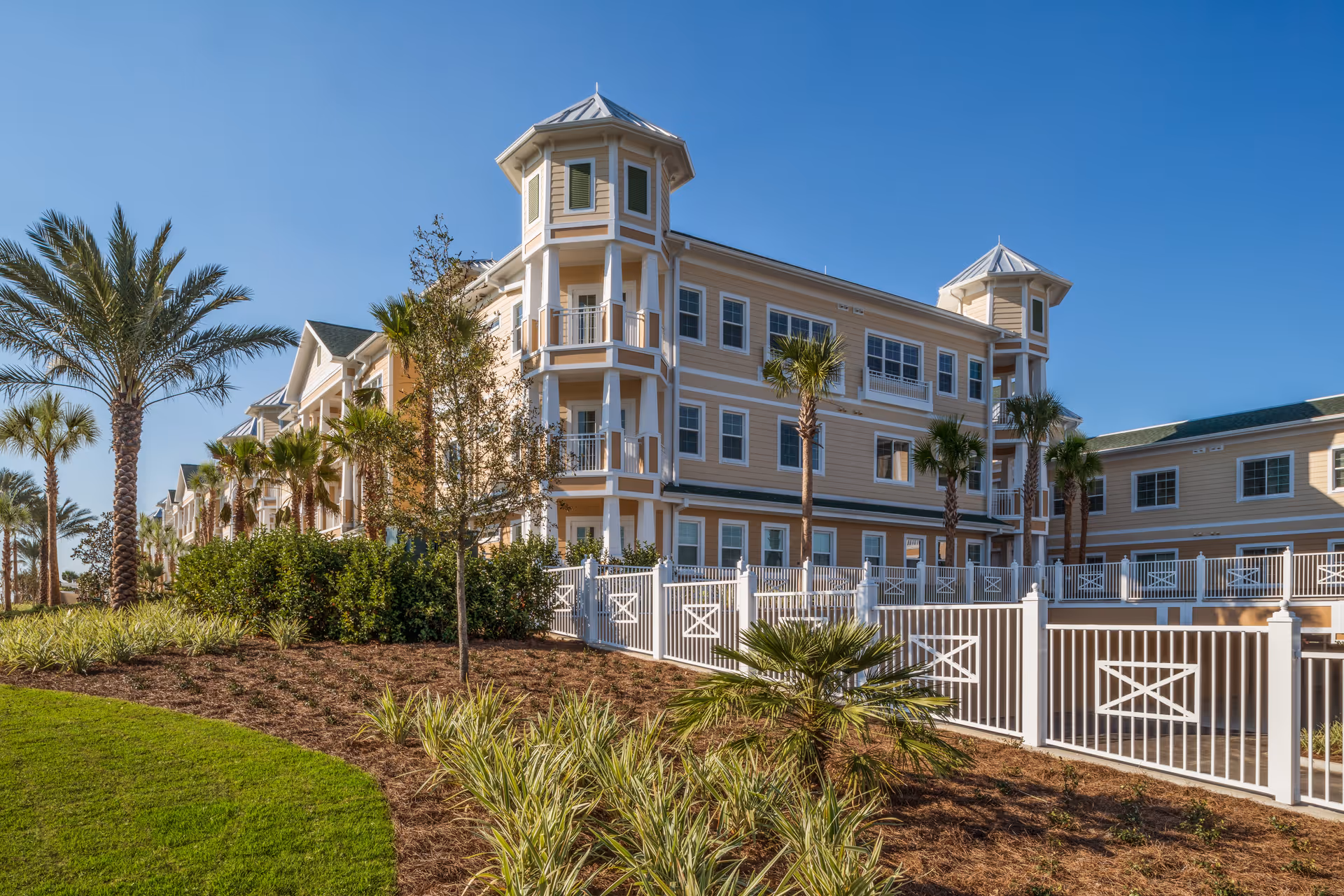 Exterior front of a multi-story pale yellow senior living building with balconies, palm trees, and white fencing under a clear blue sky.