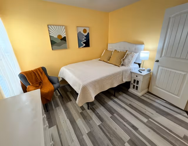 Yellow-walled bedroom featuring a white bed, nightstand, chair with an orange throw, and wall art.