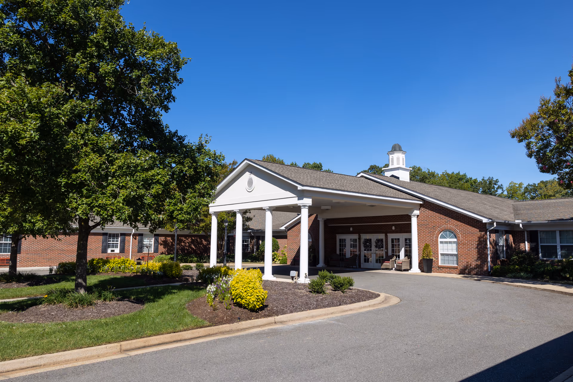 Brick senior living facility with a white portico entrance, surrounding landscaping, and blue sky.