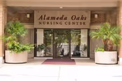 Entrance of Alameda Oaks Nursing Center with glass doors, brick facade, red mat, and potted plants.
