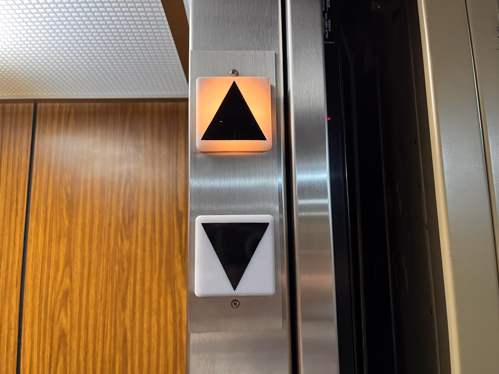 Inside an elevator, illuminated up-arrow button glows beside a dark downward button and metal walls.