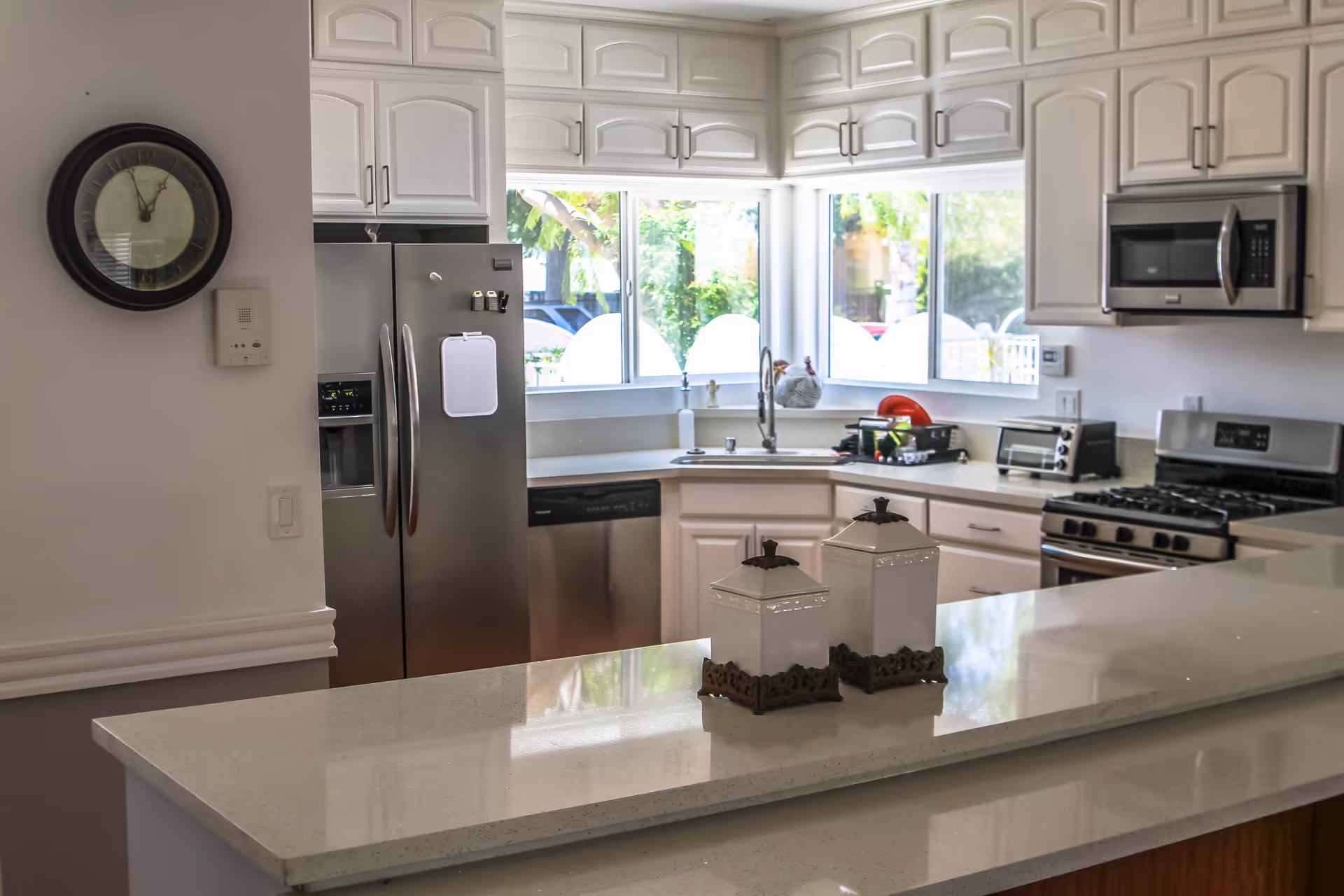 A bright kitchen with white cabinetry and stainless steel appliances including a refrigerator, dishwasher, microwave, and gas stove. There is a large window above the sink letting in natural light. On the countertop in the foreground, there are two decorative white containers with ornate bases. A round wall clock is visible on the left wall.
