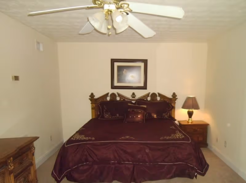 A bedroom with a large bed covered in dark purple bedding with decorative pillows. The bed has an ornate wooden headboard. There is a wooden nightstand with a lamp on the right side of the bed and a wooden dresser on the left side of the room. A ceiling fan with lights is mounted on the ceiling, and a framed picture hangs on the wall above the bed.
