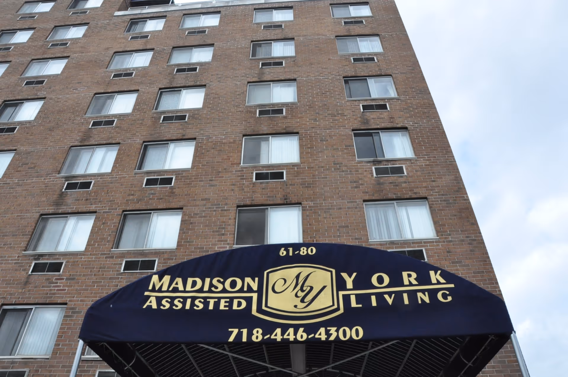 View looking up at a multi-story brick building with many windows. In the foreground, there is a blue awning with gold text that reads 'Madison York Assisted Living' along with a phone number and address number 61-80.