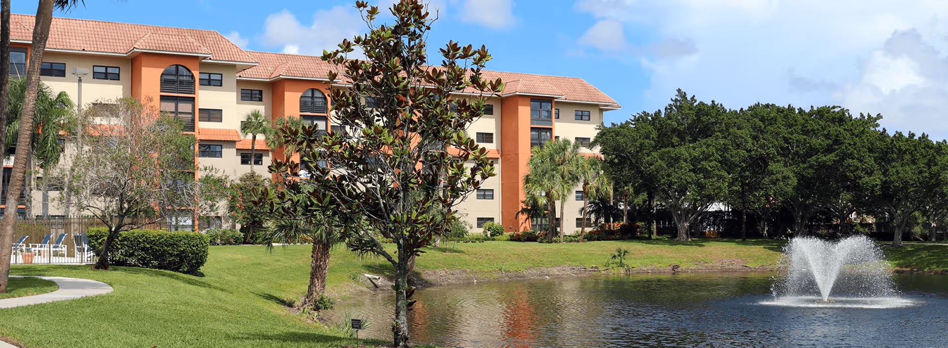 A multi-story stucco building beside a landscaped pond with a fountain, trees, and a walking path.