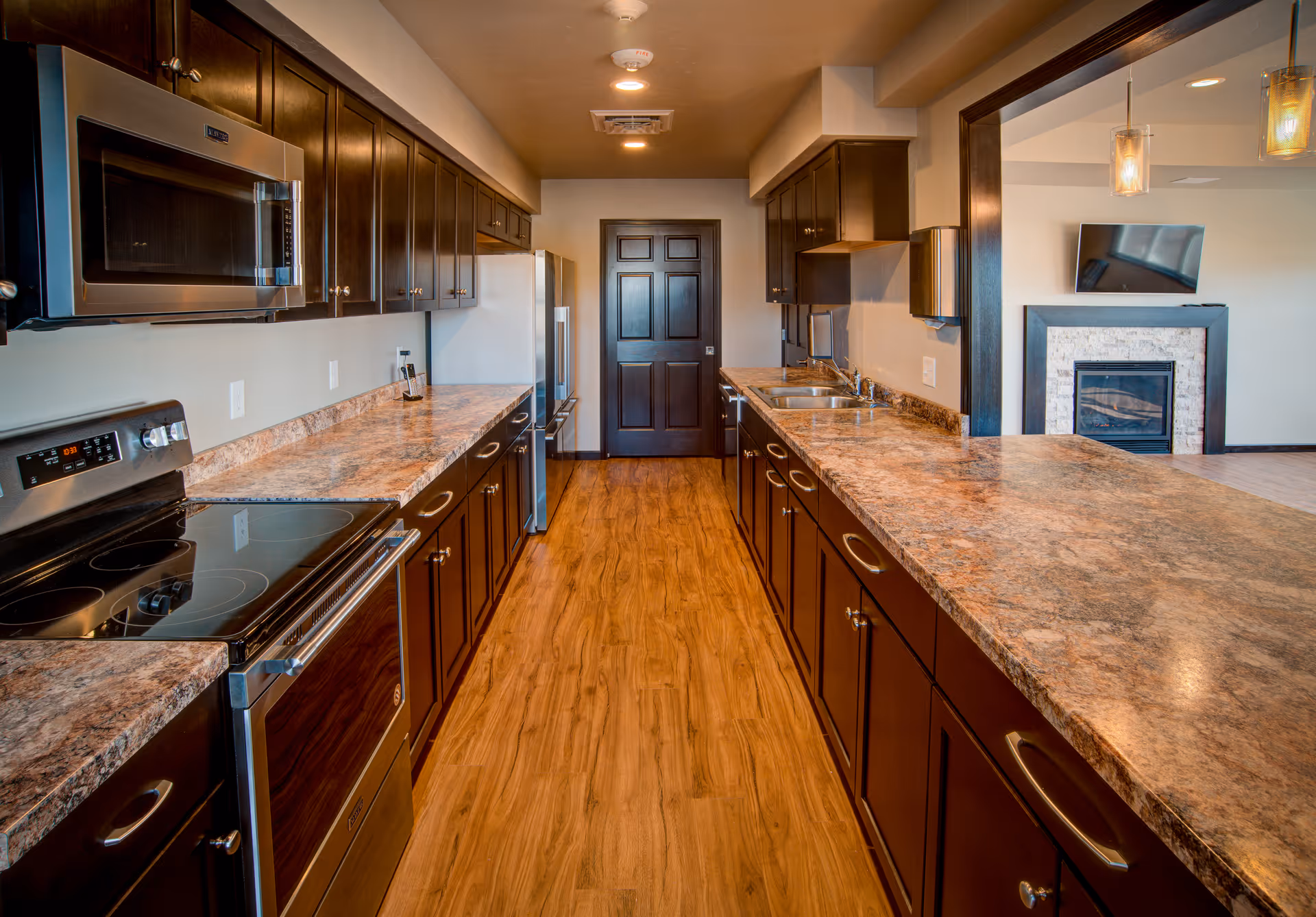A modern kitchen with dark wood cabinets, granite countertops, and stainless steel appliances including a microwave, stove, and refrigerator. The floor is wood, and the kitchen opens to a living area with a fireplace and a wall-mounted TV.