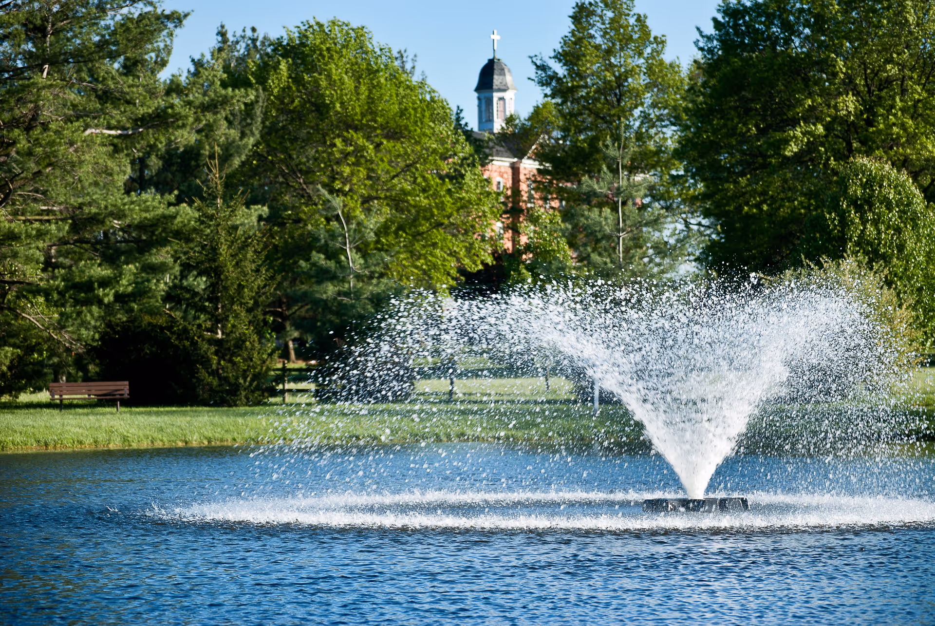 A water fountain spraying water in a pond surrounded by green trees and grass, with a building topped by a cupola and cross visible in the background.