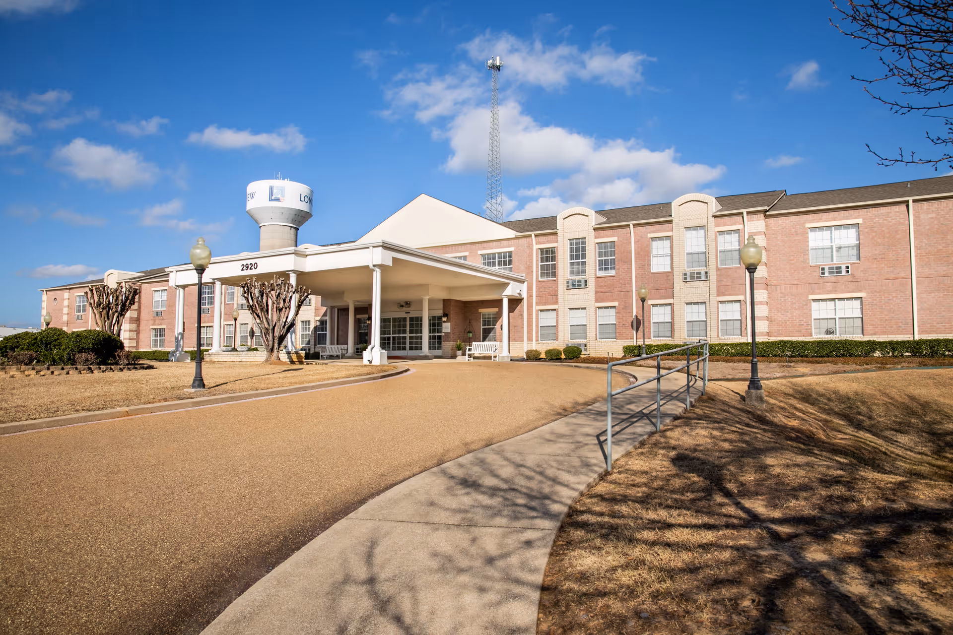 Exterior view of a senior living facility building with a covered entrance, benches, lamp posts, and a water tower in the background under a blue sky with some clouds.
