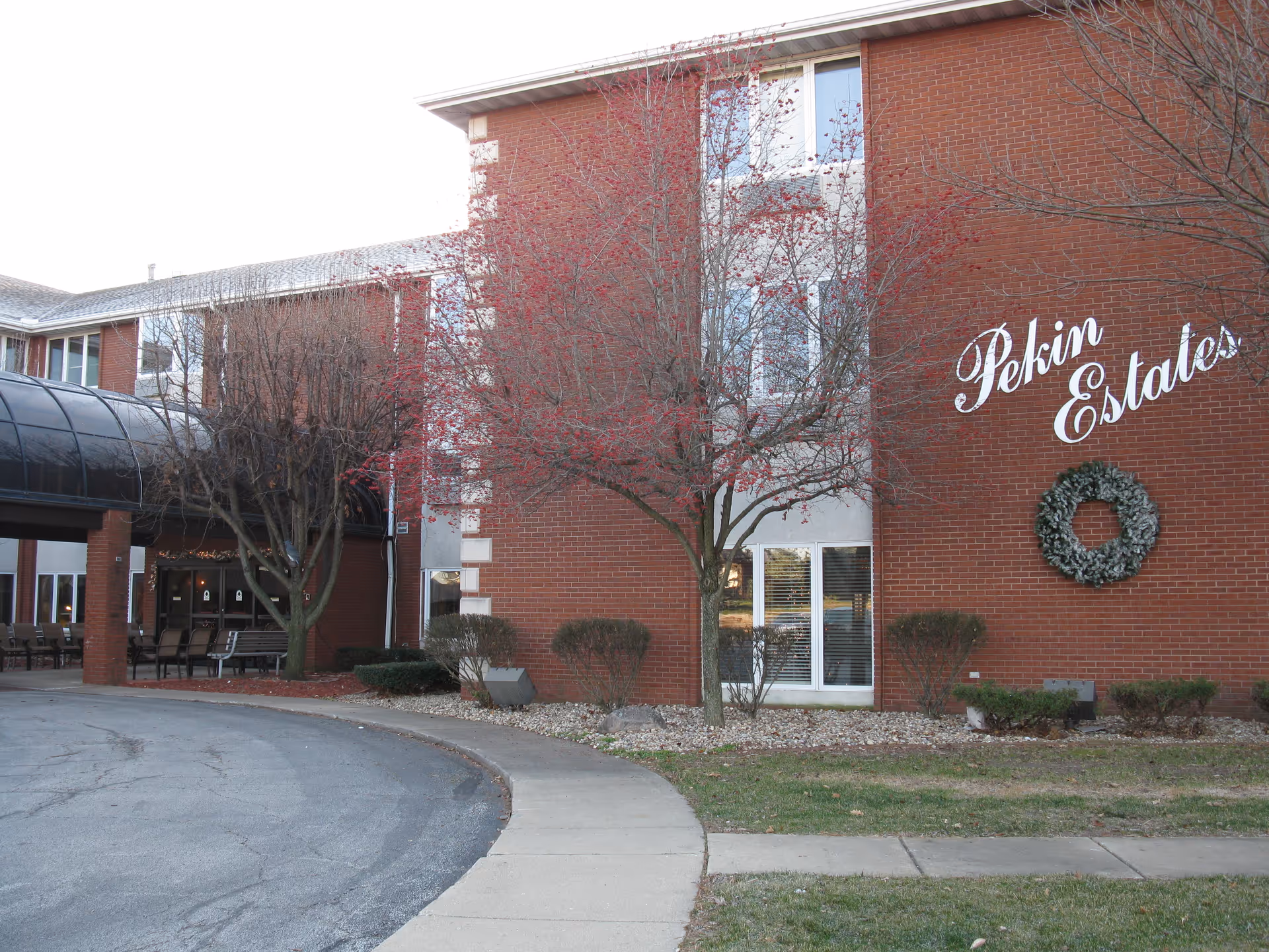 Brick senior living facility exterior with curved driveway, trees, and Pekin Estates sign.