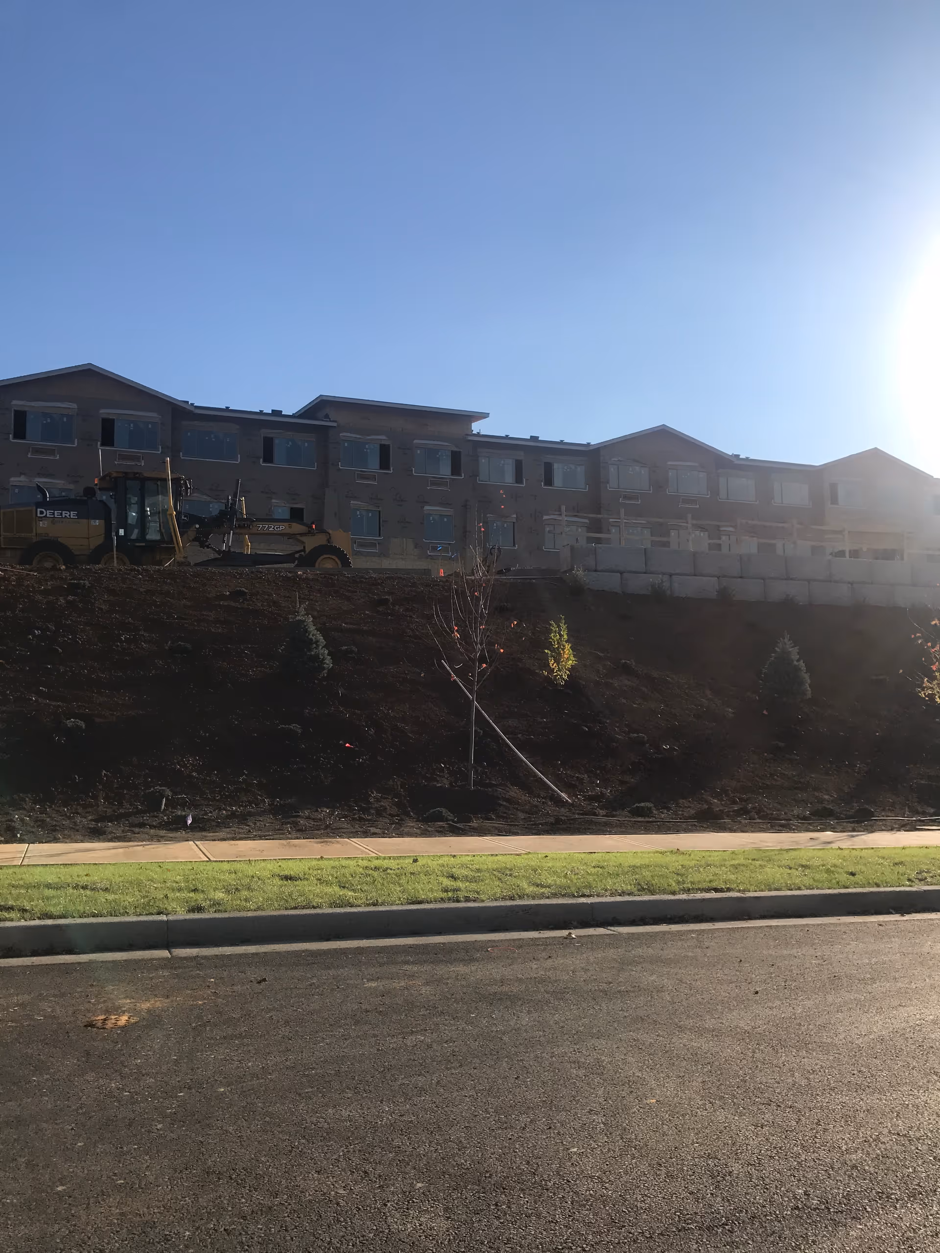 Exterior view of a multi-story building under construction on a sunny day, with construction equipment and a sloped dirt area with small trees in front.