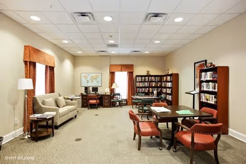 A well-lit common room with beige walls and carpeted floor featuring a beige sofa, side table with a lamp, a desk with a computer and chair, bookshelves filled with books, and tables with red cushioned chairs arranged for reading or activities.
