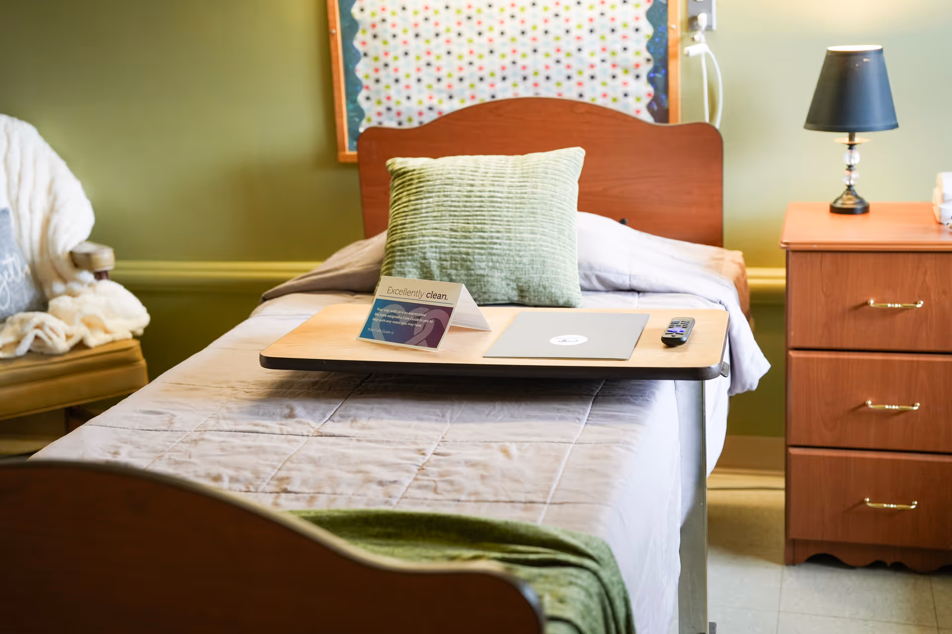 A neatly made single bed in a senior living facility room with a green pillow, a beige blanket, and a wooden overbed table holding a laptop, a remote control, and a small sign that reads 'Excellently clean.' To the right of the bed is a wooden nightstand with three drawers and a black lamp on top. To the left is a chair with a white blanket draped over it. The walls are painted green and there is a colorful bulletin board above the bed.