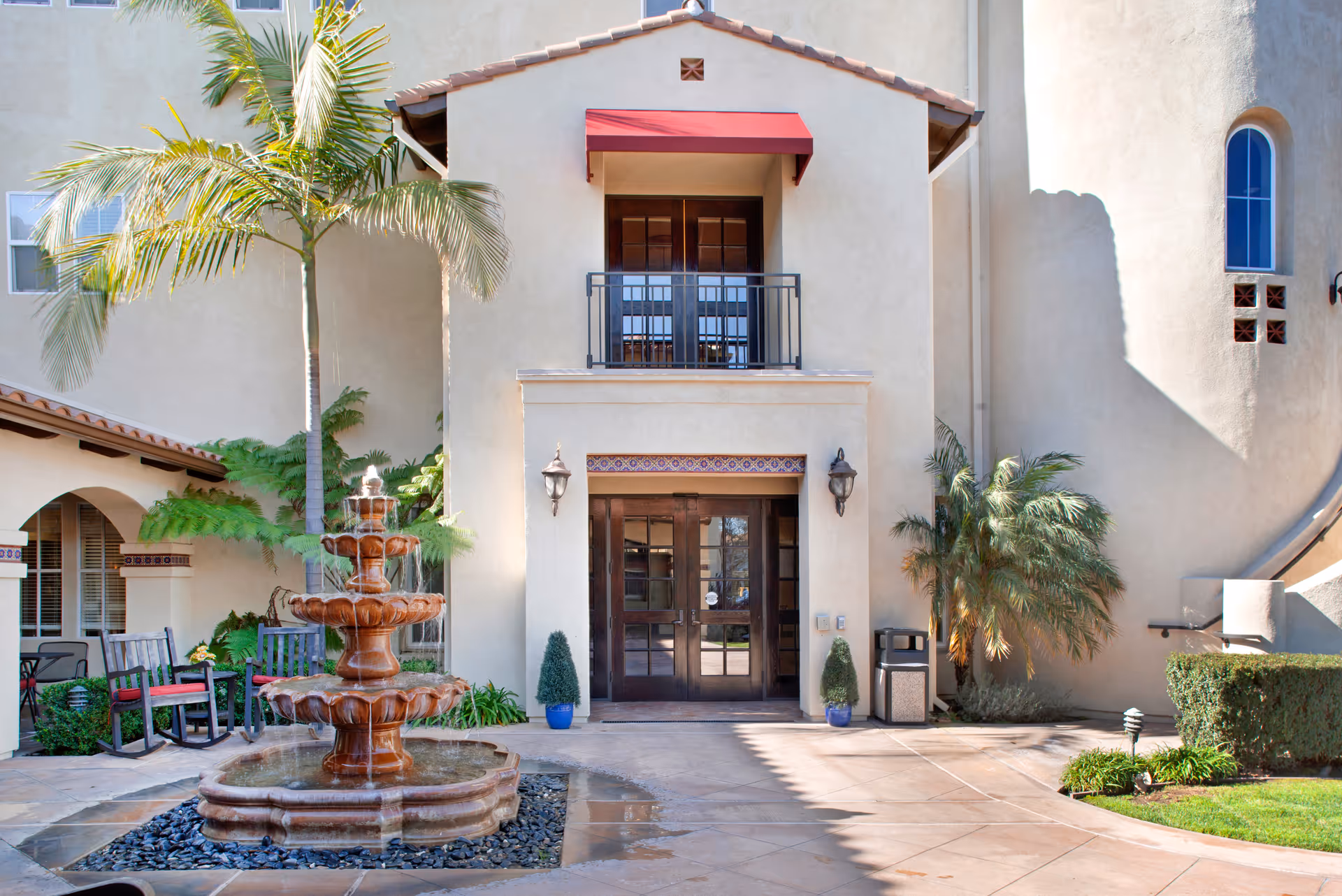 Entrance of a senior living facility with a three-tiered water fountain in front, palm trees, potted plants, and a balcony above the double glass doors. There are outdoor chairs and a small table to the left under an archway.