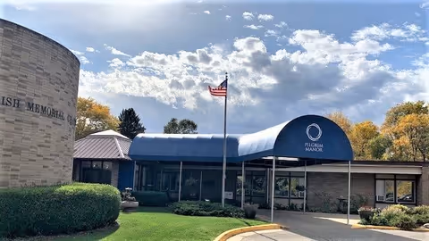 Exterior view of a single-story building with a blue awning labeled 'Pilgrim Manor' over the entrance. An American flag is flying on a flagpole in front of the building. The sky is partly cloudy and there are trees with autumn foliage in the background.