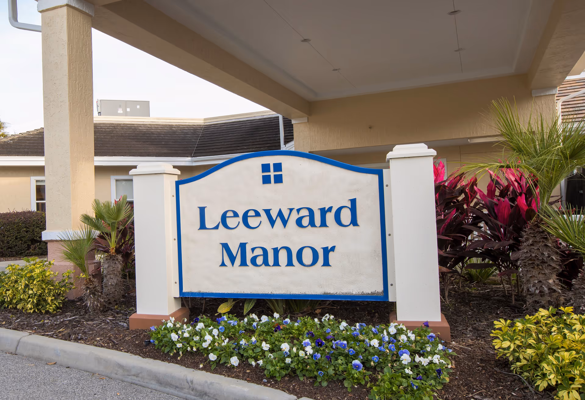 Outdoor sign reading Leeward Manor at the entrance of a beige building with tropical plants