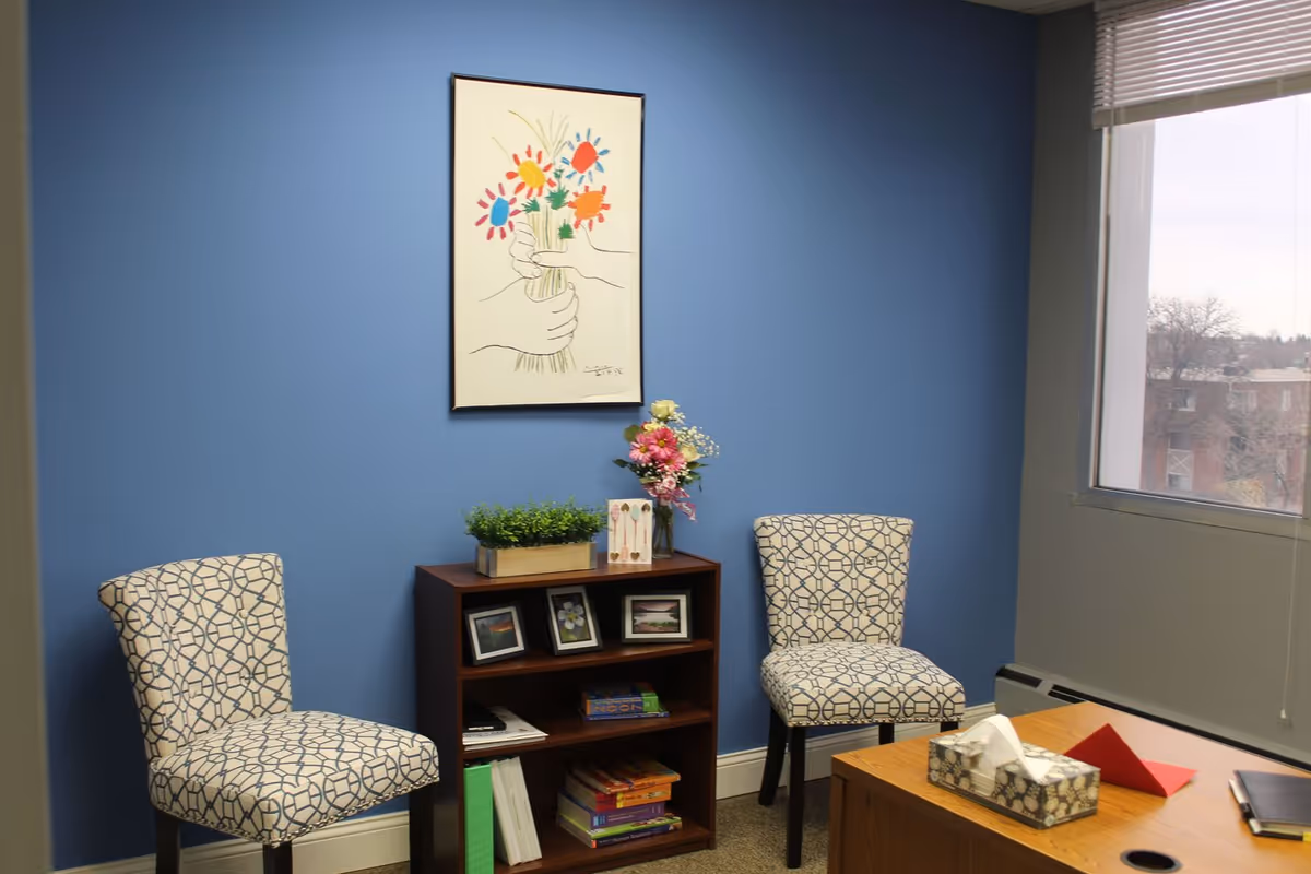 Blue-walled office lounge with two patterned chairs, a small shelf, flowers, and a window.