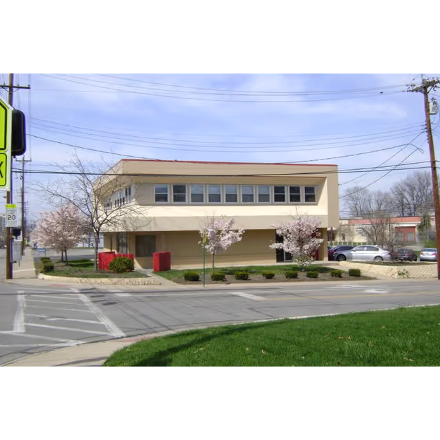 Two-story beige building on a street with blossoming trees and a manicured front lawn.