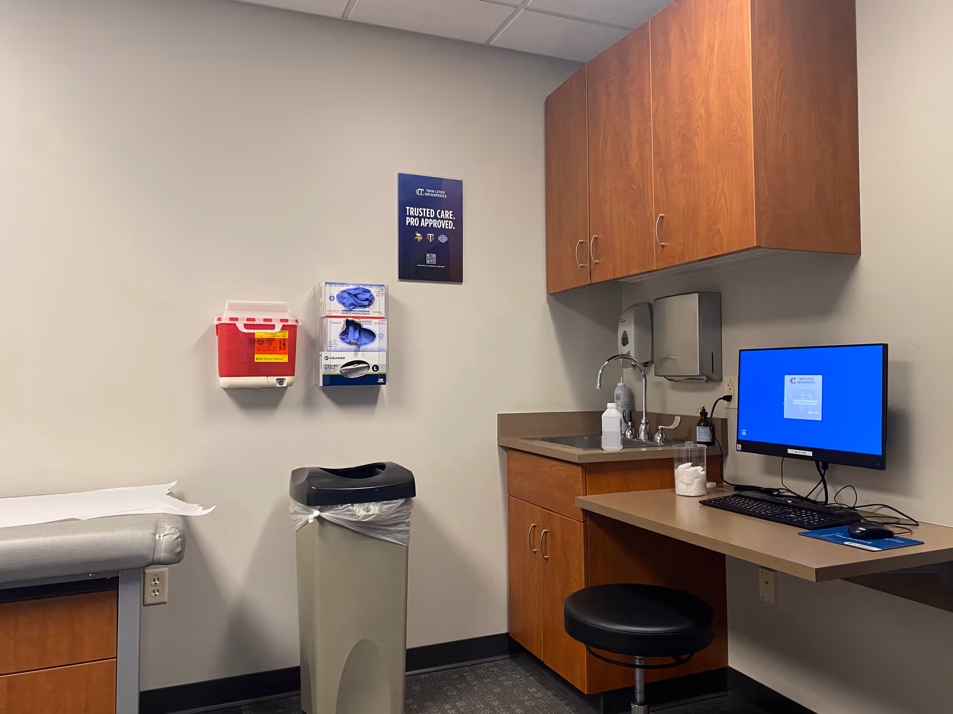 Medical examination room with a desk, computer, sink, cabinets, and exam table.