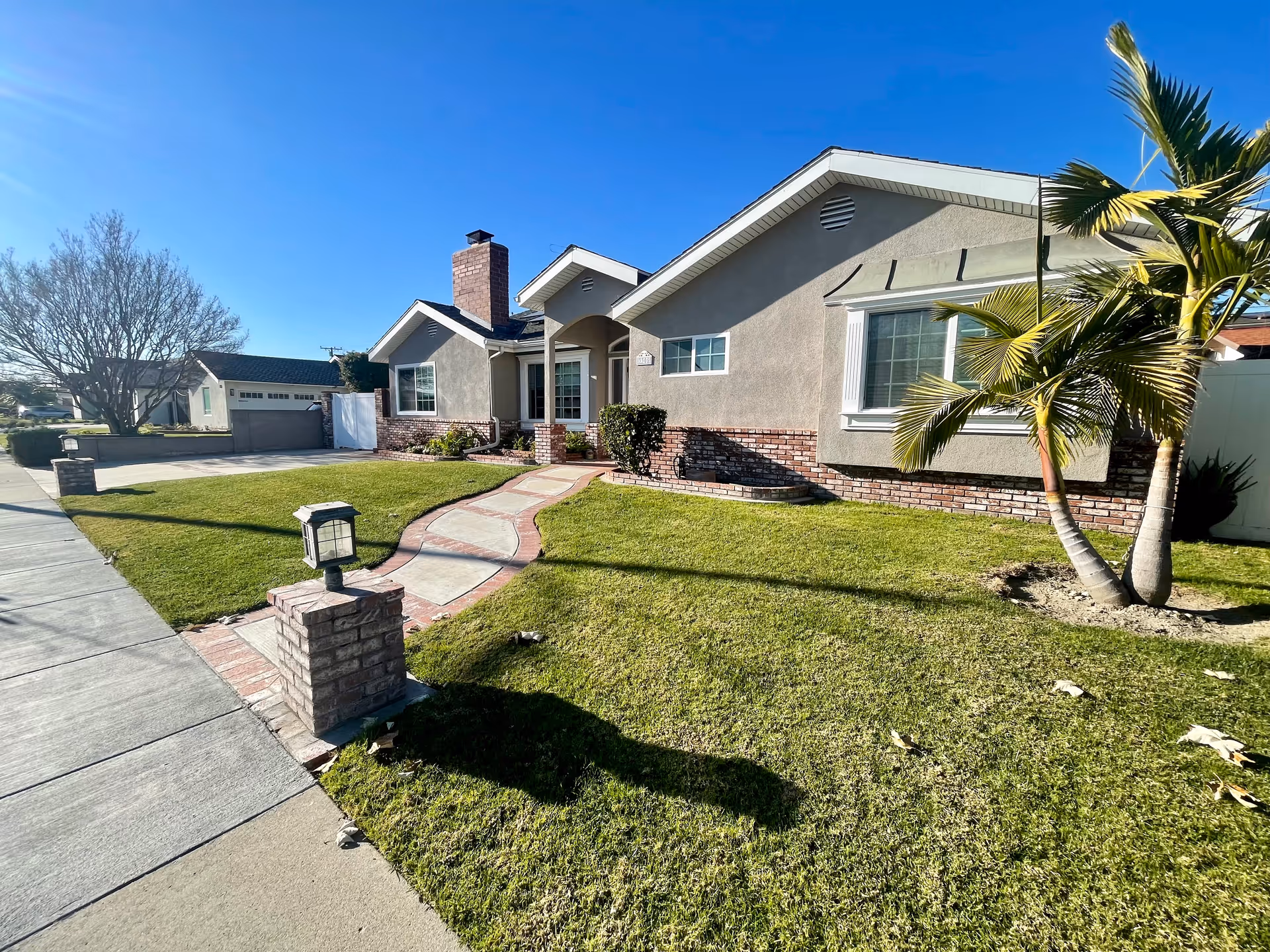 Single-story beige house with brick accents, a palm tree, curved brick walkway, and manicured lawn.