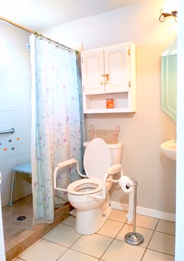 A bathroom with a white toilet equipped with safety armrests, a standing toilet paper holder, a shower area with a floral shower curtain and a built-in bench, a white wall cabinet with a small orange candle inside, and a wall-mounted sink with a mirror above it.