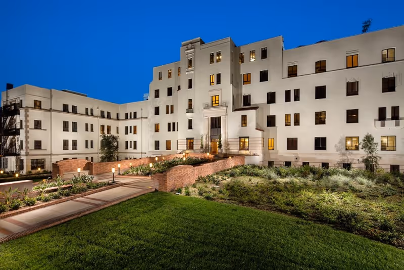 Evening exterior of Hollenbeck Terrace, a white multi-story senior living building with landscaped courtyard.